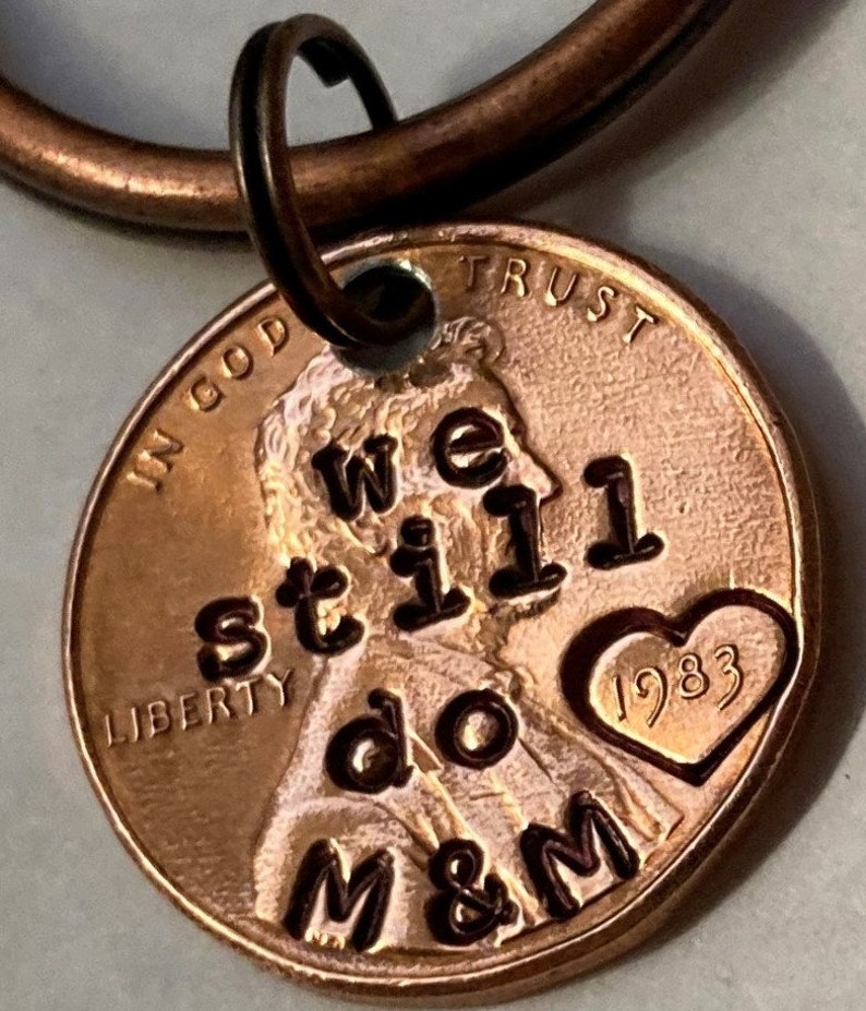 May include: A copper penny keychain with the words "We still do M&M" and a heart with the year 1983 engraved on it. The penny is attached to a copper key ring.