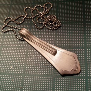 May include: A silver spoon necklace with a beaded chain. The spoon has been bent into a teardrop shape and has decorative lines on the handle.