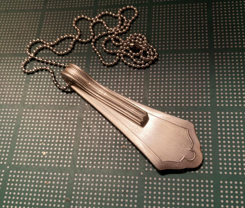 May include: A silver spoon necklace with a beaded chain. The spoon has been bent into a teardrop shape and has decorative lines on the handle.