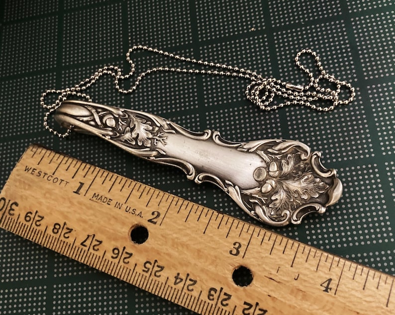May include: A silver spoon necklace with an oak leaf design. The spoon is about 6 inches long and is attached to a silver ball chain.