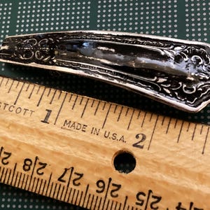 May include: A silver-colored, ornate, spoon-shaped hair clip measuring approximately 1 inch in length. The clip is lying on a wooden ruler with markings in inches.