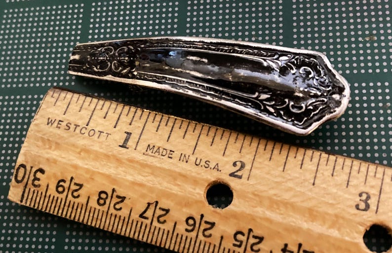 May include: A silver-colored, ornate, spoon-shaped hair clip measuring approximately 1 inch in length. The clip is lying on a wooden ruler with markings in inches.