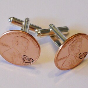 May include: A pair of cufflinks made from copper pennies with a heart and the year 2012 engraved on each. The cufflinks are attached to silver-toned metal bars.