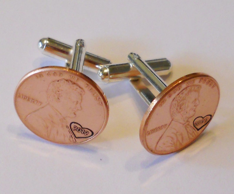 May include: A pair of cufflinks made from copper pennies with a heart and the year 2012 engraved on each. The cufflinks are attached to silver-toned metal bars.