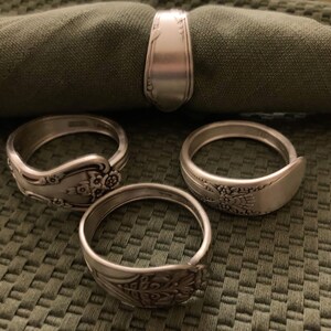 May include: Four silver napkin rings made from bent silverware with ornate designs. The rings are on a green fabric background.