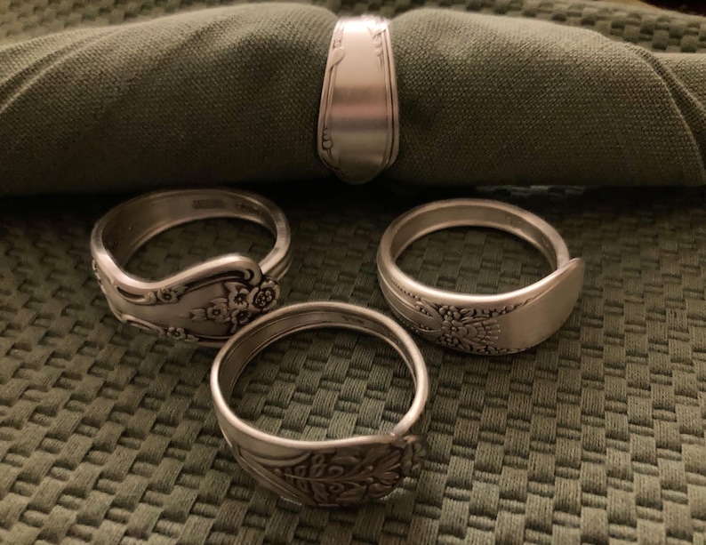 May include: Four silver napkin rings made from bent silverware with ornate designs. The rings are on a green fabric background.