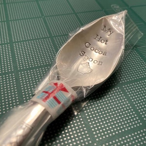 May include: A silver spoon with the words "My Hot Cocoa Spoon" engraved on it. The spoon is wrapped in clear plastic with a red and white striped ribbon.