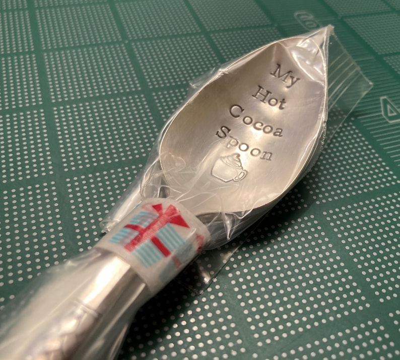 May include: A silver spoon with the words "My Hot Cocoa Spoon" engraved on it. The spoon is wrapped in clear plastic with a red and white striped ribbon.