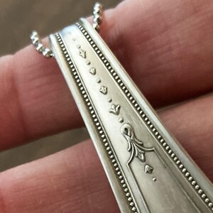 May include: Close-up of a silver spoon handle with a decorative design of small flowers and a larger floral motif. The spoon handle is part of a necklace.