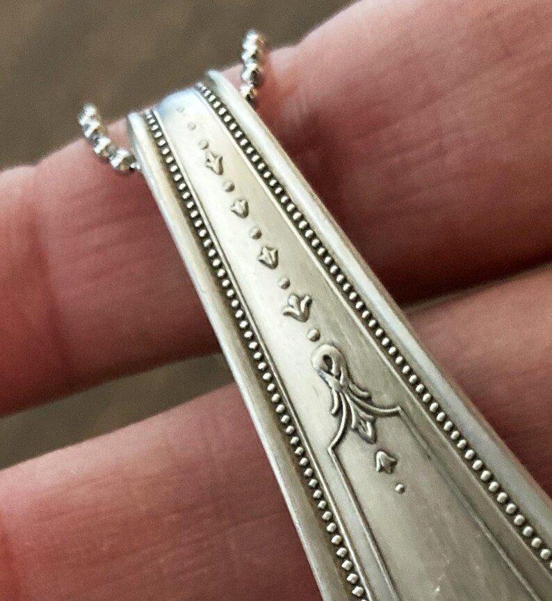May include: Close-up of a silver spoon handle with a decorative design of small flowers and a larger floral motif. The spoon handle is part of a necklace.