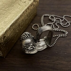 May include: A silver spoon necklace with a detailed grape design. The spoon is bent into a heart shape and hangs from a silver ball chain.