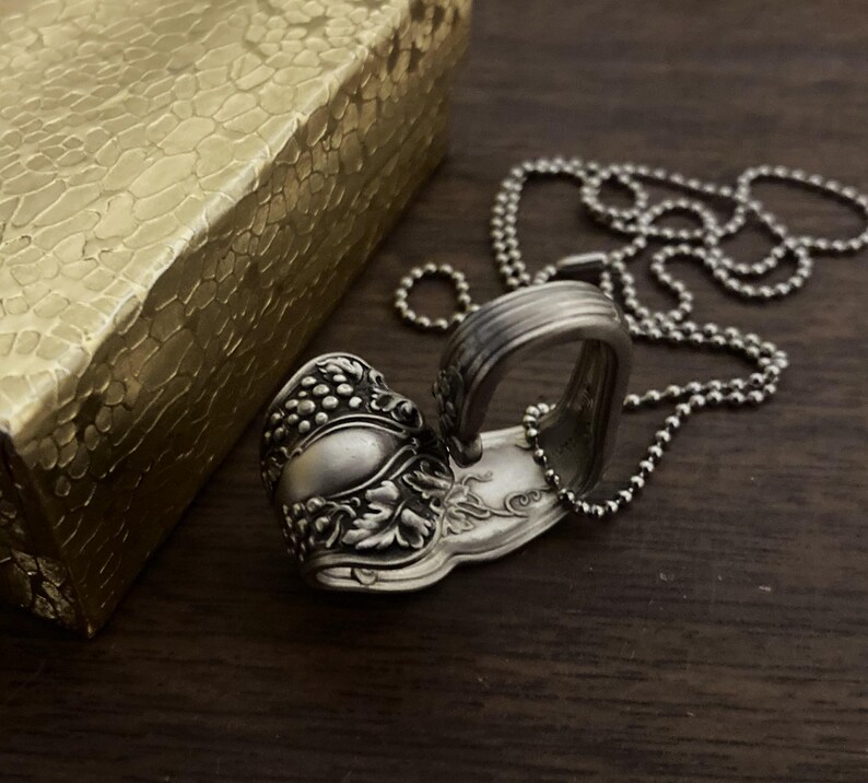 May include: A silver spoon necklace with a detailed grape design. The spoon is bent into a heart shape and hangs from a silver ball chain.