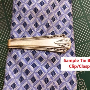 May include: A silver tie bar with an Art Deco design rests on a blue and white patterned tie. The tie bar is rectangular with a decorative end. The tie has a diamond pattern.