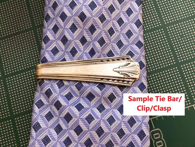 May include: A silver tie bar with an Art Deco design rests on a blue and white patterned tie. The tie bar is rectangular with a decorative end. The tie has a diamond pattern.