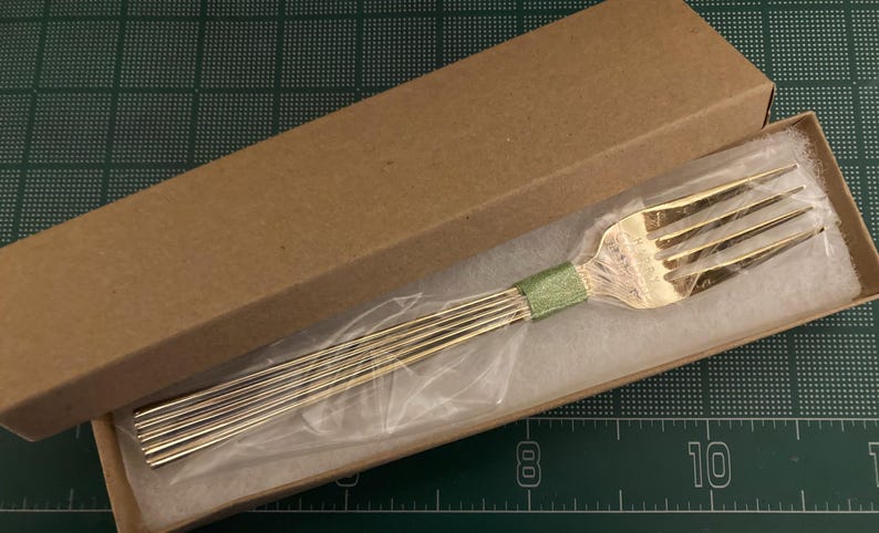 May include: A silver fork with a green ribbon wrapped around the handle is displayed in a brown cardboard box with white tissue paper.