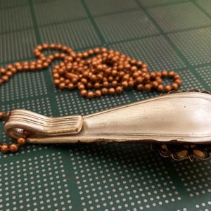 May include: A silver spoon necklace with a copper chain. The spoon is bent into a curved shape and has a decorative design on the back.