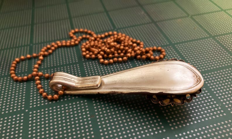 May include: A silver spoon necklace with a copper chain. The spoon is bent into a curved shape and has a decorative design on the back.
