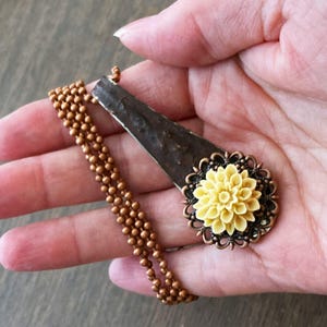 May include: A copper chain necklace with a pendant featuring a yellow flower with a brown metal frame. The pendant is made from a repurposed metal spoon.