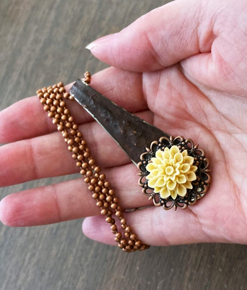 May include: A copper chain necklace with a pendant featuring a yellow flower with a brown metal frame. The pendant is made from a repurposed metal spoon.