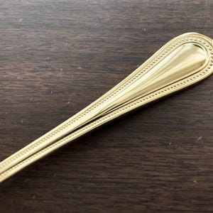 May include: Close-up of a gold-colored spoon with a decorative beaded border. The spoon's handle has a teardrop shape and a polished finish. The spoon is set against a dark wood grain surface.