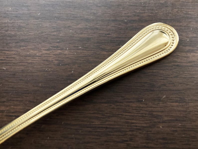 May include: Close-up of a gold-colored spoon with a decorative beaded border. The spoon's handle has a teardrop shape and a polished finish. The spoon is set against a dark wood grain surface.