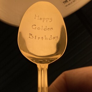 May include: A gold-colored spoon with the words "Happy Golden Birthday" engraved on the bowl. The spoon has a decorative handle and is set against a dark background.
