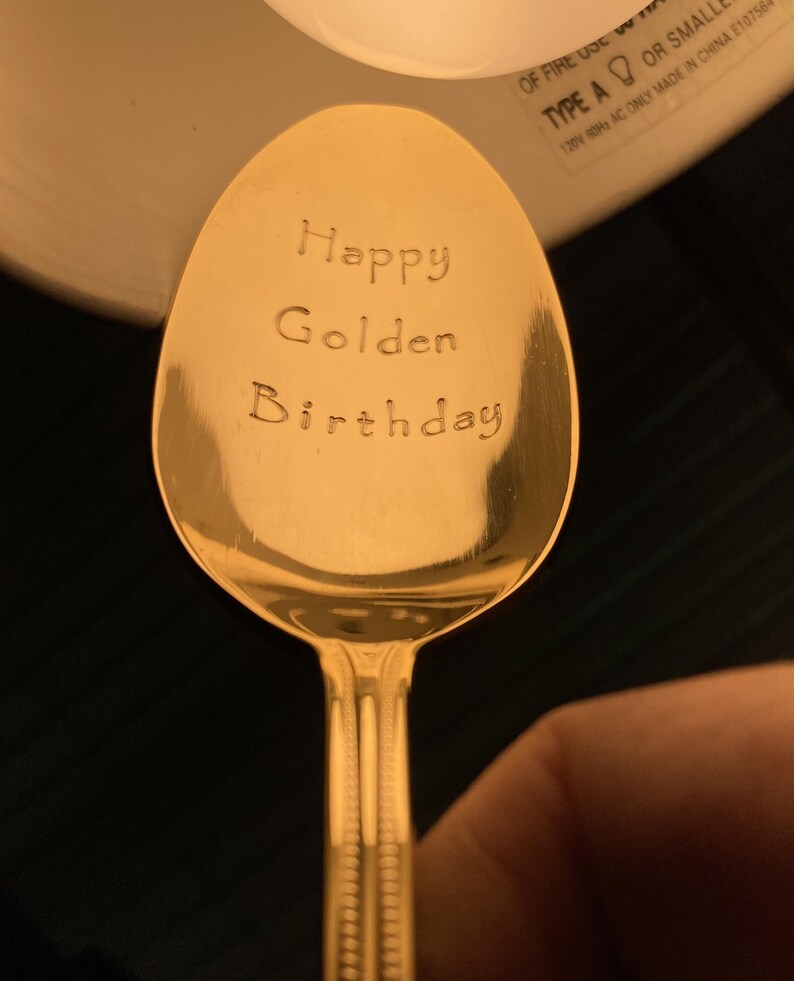 May include: A gold-colored spoon with the words "Happy Golden Birthday" engraved on the bowl. The spoon has a decorative handle and is set against a dark background.