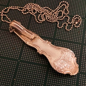 Puede incluir: Un collar de cuchara de plata con un dise&ntilde;o floral en el mango. La cuchara est&aacute; unida a una cadena de plata.