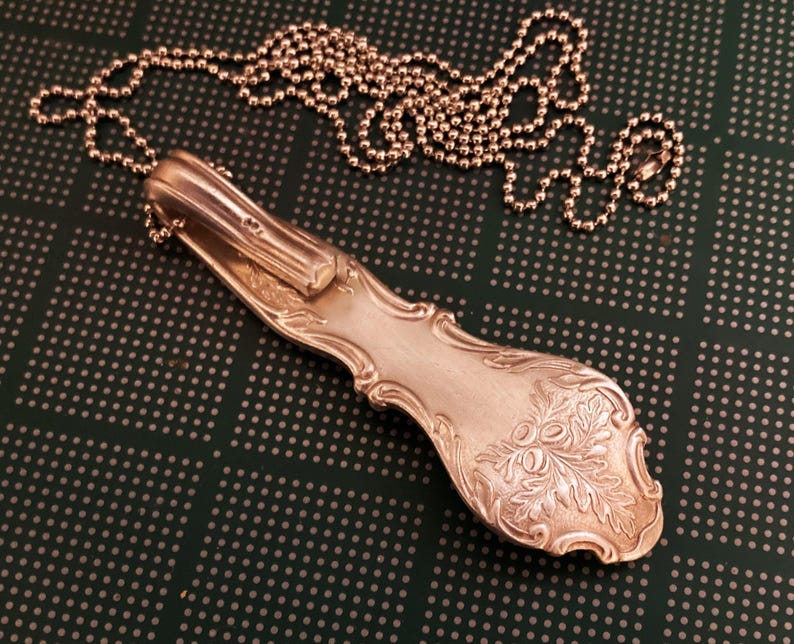 Puede incluir: Un collar de cuchara de plata con un dise&ntilde;o floral en el mango. La cuchara est&aacute; unida a una cadena de plata.