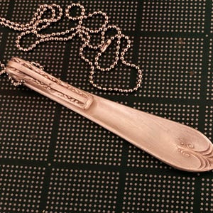 Puede incluir: Un collar de cuchara de plata con un dise&ntilde;o decorativo en el mango. La cuchara est&aacute; unida a una cadena de plata.