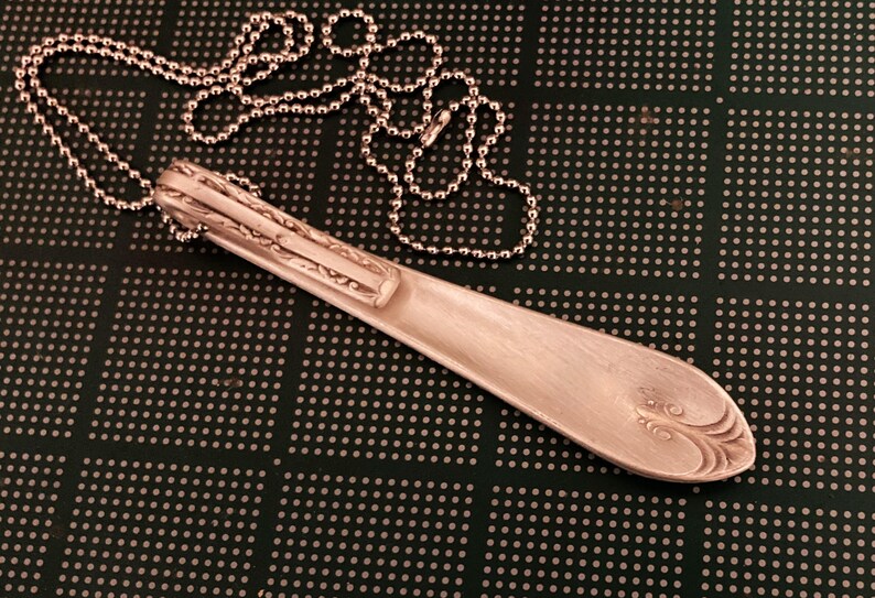 Puede incluir: Un collar de cuchara de plata con un dise&ntilde;o decorativo en el mango. La cuchara est&aacute; unida a una cadena de plata.