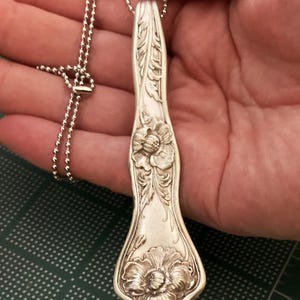 May include: A silver spoon necklace with a floral design. The spoon is engraved with two large flowers and intricate details. The necklace is made with a silver ball chain.