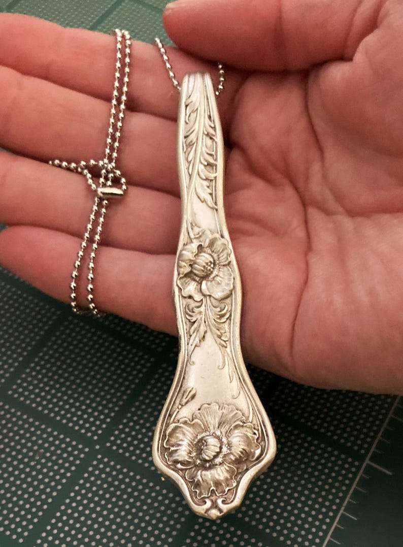 May include: A silver spoon necklace with a floral design. The spoon is engraved with two large flowers and intricate details. The necklace is made with a silver ball chain.