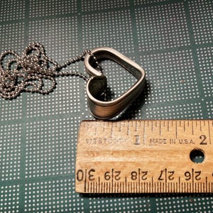 May include: A silver heart-shaped pendant necklace with a chain. The pendant is made from a spoon and is about 1.5 inches in length. The necklace is on a green surface with a ruler for scale.