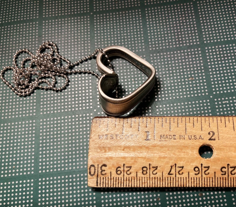 May include: A silver heart-shaped pendant necklace with a chain. The pendant is made from a spoon and is about 1.5 inches in length. The necklace is on a green surface with a ruler for scale.