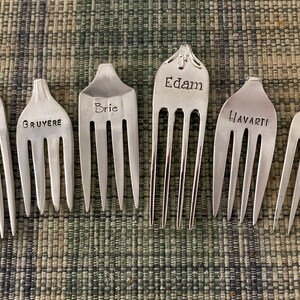 May include: Seven silver forks with different cheese names engraved on them: Goat, Gruyere, Brie, Edam, Havarti, and Bleu.