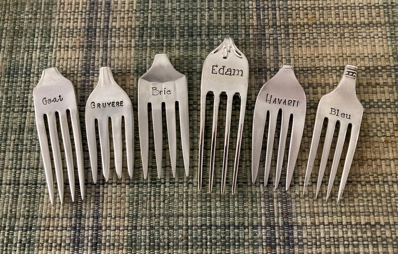 May include: Seven silver forks with different cheese names engraved on them: Goat, Gruyere, Brie, Edam, Havarti, and Bleu.
