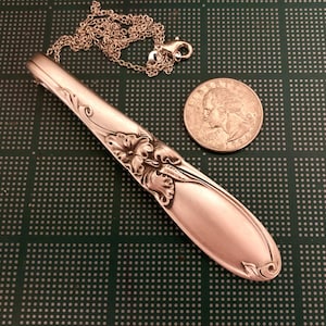 Puede incluir: Un collar con colgante de plata hecho de una cuchara, con detalles florales y de volutas. El colgante est&aacute; unido a una cadena fina con un cierre. Una moneda est&aacute; al lado del colgante para la comparaci&oacute;n de tama&ntilde;o.