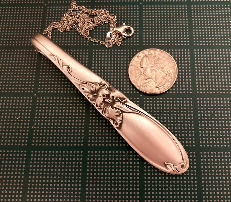 Puede incluir: Un collar con colgante de plata hecho de una cuchara, con detalles florales y de volutas. El colgante est&aacute; unido a una cadena fina con un cierre. Una moneda est&aacute; al lado del colgante para la comparaci&oacute;n de tama&ntilde;o.