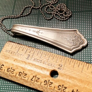 May include: A silver spoon necklace with an ornate design and a ball chain. The spoon is about 2 inches long and the chain is about 18 inches long.