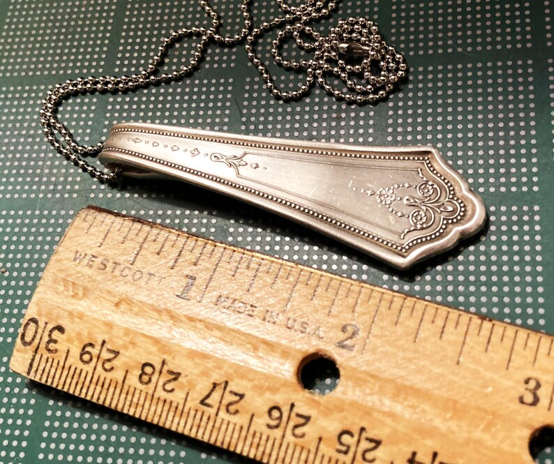 May include: A silver spoon necklace with an ornate design and a ball chain. The spoon is about 2 inches long and the chain is about 18 inches long.