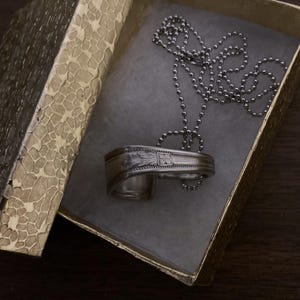 May include: A silver spoon necklace with a decorative design, in a gold box with a textured pattern. The necklace is on a silver ball chain.