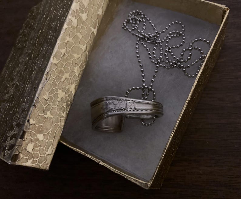 May include: A silver spoon necklace with a decorative design, in a gold box with a textured pattern. The necklace is on a silver ball chain.