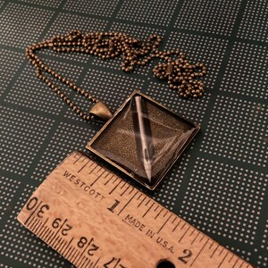 K&ouml;nnte beinhalten: Eine bronzene Halskette mit einem quadratischen Anh&auml;nger, der eine silberne Nadel in Harz gefasst zeigt. Der Anh&auml;nger ist etwa 2,5 cm gro&szlig;.