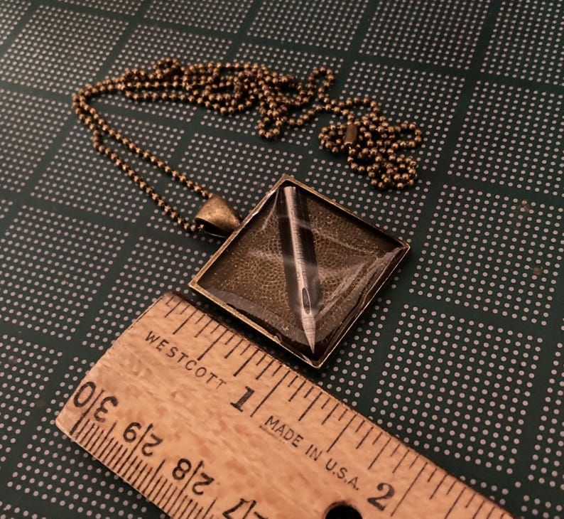 K&ouml;nnte beinhalten: Eine bronzene Halskette mit einem quadratischen Anh&auml;nger, der eine silberne Nadel in Harz gefasst zeigt. Der Anh&auml;nger ist etwa 2,5 cm gro&szlig;.