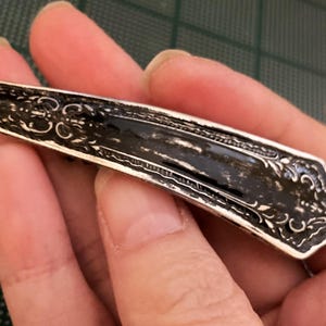 May include: A silver hair clip with an ornate design. The clip is black and has a shiny finish.