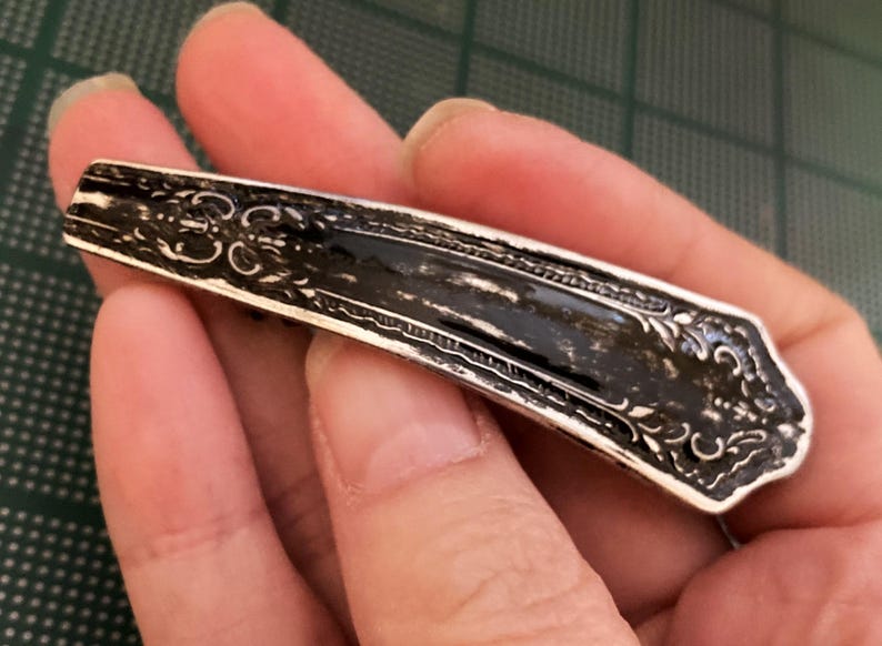 May include: A silver hair clip with an ornate design. The clip is black and has a shiny finish.