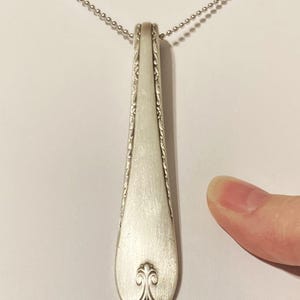 Puede incluir: Un collar con un colgante de cuchara de plata con un dise&ntilde;o adornado en el mango. La cuchara est&aacute; unida a una cadena de plata.