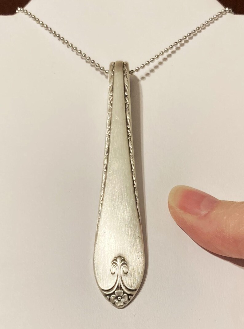 Puede incluir: Un collar con un colgante de cuchara de plata con un dise&ntilde;o adornado en el mango. La cuchara est&aacute; unida a una cadena de plata.