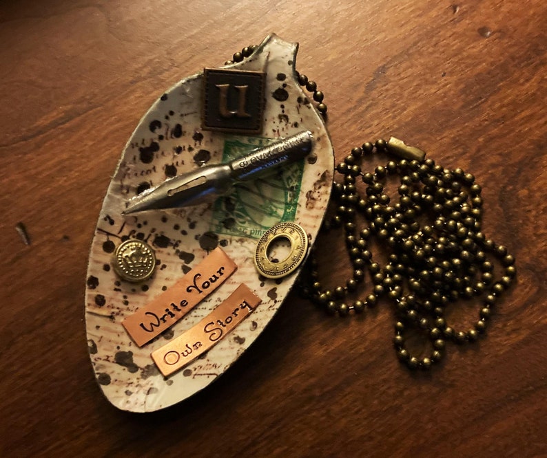 Op de afbeelding: Een wit keramisch gerecht in de vorm van een blad met een bruin, zwart en gouden ontwerp. Het gerecht heeft een metalen penpunt, een gouden knoop en twee koperen labels met de tekst "Write Your Own Story". Er ligt een gouden ketting naast het gerecht.